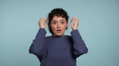 Close up young curly woman in blue sweater shows an explosion gesture near the temples. Portrait beautiful female standing on isolated light blue background. People emotions lifestyle concept