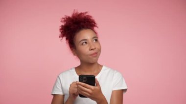 Smiling African American redhead curly woman using smartphone typing browsing thinking about buying in online store. Female holding mobile phone makes decision about online purchase on pink background