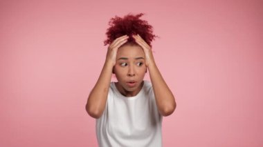 Portrait shocked African American redhead curly woman looking camera puts hands on head. Excited female in white t-shirt standing on isolated pink background with copy space. People emotions lifestyle