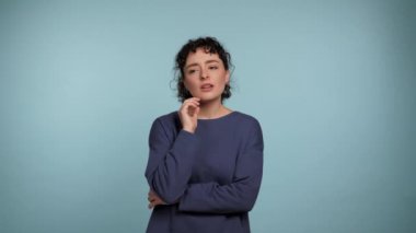 Pensive young curly woman in blue sweater posing looking around, thinking raising finger with idea. Thoughtful beautiful female thinks how to solve the problem on isolated light blue background