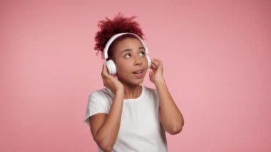 Neşeli gülümseyen Afro-Amerikan kıvırcık saçlı kadın kulaklık danslarında müzik dinliyor. Mutlu kadın portresi. İzole edilmiş pembe arka planda beyaz tişört giyiyor.