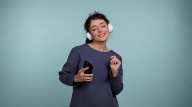 Portre gülümseyen kıvırcık kadın akıllı telefonu elinde tutuyor kulaklıkla müzik dinliyor, dans ediyor. İzole edilmiş, açık mavi arka planda, fotokopi alanı olan mutlu, güzel bir kadın. İnsanların duyguları