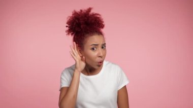 Kafası karışmış Afro-Amerikan kıvırcık saçlı, eli kulağında bir kadın, söylenenleri duyamıyor. İzole edilmiş pembe arka planda duran beyaz tişört giyen, fotokopi alanı olan kadın.