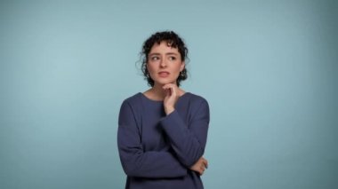Pensive young curly woman in blue sweater posing looking around, thinking raising finger with idea. Thoughtful beautiful female thinks how to solve the problem on isolated light blue background