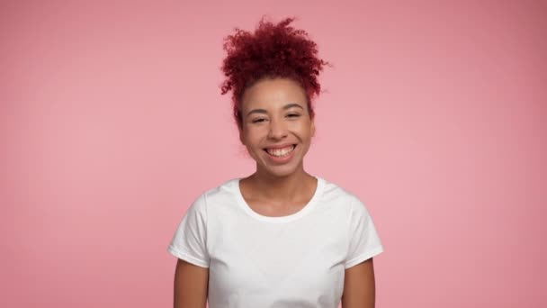 笑得很开心的年轻的非洲裔美国女人 红头发卷曲 身穿白色T恤 摆出一副相机的样子 站在孤立的粉色背景上的正面女性的画像 人的情感生活方式 — 图库视频影像