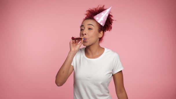 快乐的非裔美国红头发卷曲女人头戴生日帽吹口哨庆祝节日 快乐而积极的女性 穿着白色T恤衫 背景是粉红色的 有复制空间 — 图库视频影像