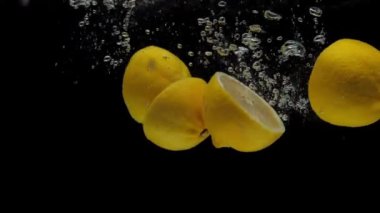 Yavaş çekim sarı limon yarımları siyah arkaplanda şeffaf suya dökülüyor. Akvaryuma taze narenciye sıçrıyor. Meyveler, hava kabarcıkları