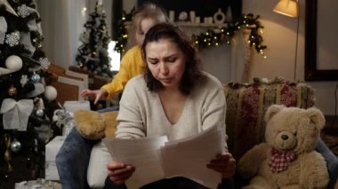 Noel ağacıyla süslenmiş bir çocukla birlikte oturma odasında oturan üzgün orta yaşlı bir kadın, kağıt mektupları okurken üzüldü. Yüksek kalite 4k görüntü