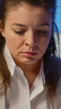 Dikey video: Aşırı çalışan iş kadınının gece geç saatlere kadar çalışan bir iş ofisinde kamera karşısında bitkin bir şekilde iç çekişi. Teknoloji ağı kablosuz bağlantısını kullanarak odaklanmış bir çalışan