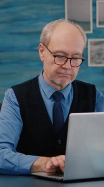 Dikey video: Emekli yönetici kameranın önünde oturmuş dizüstü bilgisayarda yazdıktan sonra gülümserken kıdemli eş arka planda bir kitap okuyor. Çevrimiçi toplantı için hazırlanmış adam