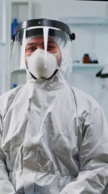 Dikey video: Donanımlı laboratuvarda koruyucu maske takarak kameraya gülümseyen deneyimli bilim adamının portresi. Aşı için yüksek teknoloji ve kimya araçları kullanılarak virüs evrimi inceleniyor