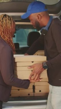 Dikey video: Erkek pizzacı kurye ofis binasındaki müşterilere pizza kutuları veriyor, araba bagajında büyük bir restoran siparişi teslim ediyor. Fast food paketlerini araçtan çıkarıp...