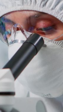 Dikey video: Laboratuvar teknisyeninin koruma giysisiyle portresi mikroskop kullanarak laboratuarda örnekler inceliyor. Çeşitli bakteri, doku ve kan örnekleriyle çalışan bilim adamları