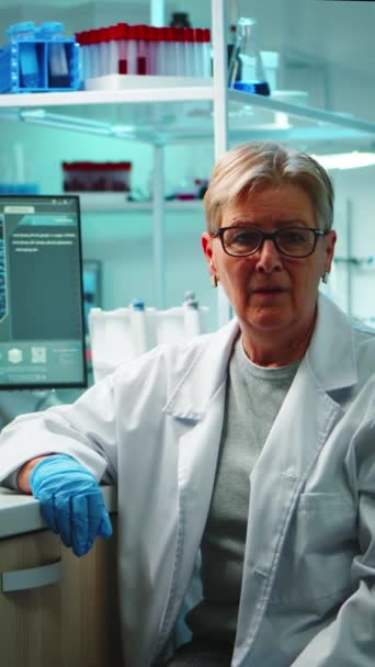 垂直ビデオ 夜遅くに設備の整った研究室でカメラの前で疲労顔を持つ古い化学者の女性の肖像画 研究のためのハイテクと化学ツールを使用してウイルス進化を調べるチーム — ストック動画