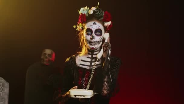 死んだ女性スタジオで固定電話を保持し コードとオフィスの電話で話をし 頭蓋骨を身に着けている 聖メキシコの儀式を祝うサンタ マエルテの衣装の女性 手持ち撮影 — ストック動画