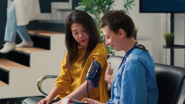 待機エリアロビーでアジアの患者と心臓病の相談を行う女性看護師 計量計で高血圧を測定 医療機器による血圧や血圧の確認 — ストック動画