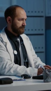 Dikey video: Hastane ofisinde gece vardiyasında çalışan yorgun doktor, bilgisayar daktilo raporunda tıbbi uzmanlığı analiz ediyor. Stresli doktor planlama hastası