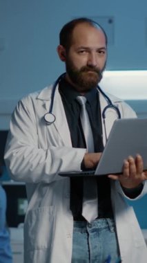 Dikey video: Tıbbi ceket giyen gülümseyen doktor portresi ve steteskop elinde dizüstü bilgisayar, sağlık hizmeti uzmanlığı yazımı. Klinik personeli hastane ofisinde saatlerce çalışıyor. İlaç.