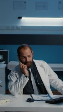 Dikey video: Genel pratisyen hekim sabit hatlı telefona cevap veriyor uzaktan doktorla tıbbi uzmanlık hakkında konuşuyor. Hastane personeli saatlerce ofiste çalışıyor. İlaç servisi.