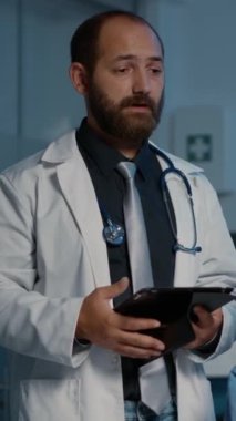 Dikey video: Doktor elinde tablet bilgisayarla hasta tanı uzmanlığı yazarken hemşirenin sağlık tedavisini tartıştığı sağlık raporunu analiz ediyor. Klinik çalışanları gece geç saatlere kadar çalışıyor.