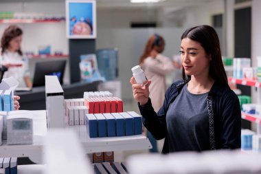 Elinde ilaç şişesi olan kadın ilaç reçetesine bakıyor. Bağışıklık sistemi için eczaneden ilaç alıyor. Vitamin dolu eczane rafları, satın alınmaya hazır takviyeler