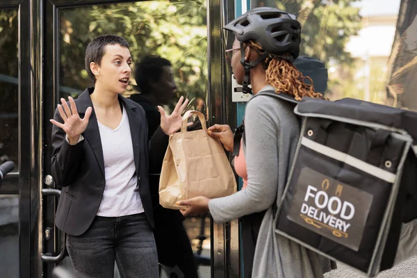 Unsatisfied client refuse taking late food order, delivery delay, courier and angry customer. Woman giving impulsive furious employee takeaway meal near office building outdoors