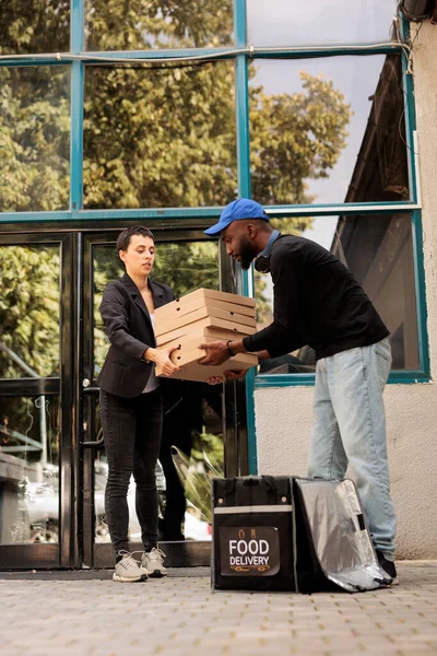 Pizzeria deliveryman passing client pizza boxes huge stack, man taking out order from thermal bag in front of office building outdoors. Food delivery service, african american courier holding meal