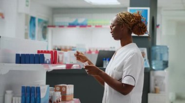 Gülümseyen Afrikalı Amerikalı eczacı eczanedeki raflarda uyuşturucuları kontrol ediyor, ilaçlarla müşterilere yardım ediyor, ilaç şişeleri ve vitamin kutuları tutuyor. Eczane masasında çalışıyorum. El kamerasıyla..