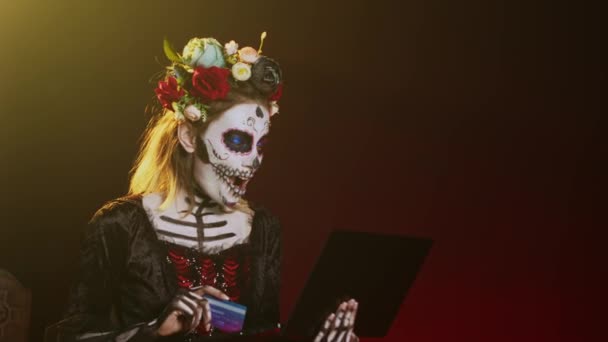 Santa Muerteはラップトップでオンラインショッピングをし スタジオでクレジットカードで支払います Pc上で電子決済取引を行う ハロウィンの伝統に死の神聖な女神のように見えます 手持ち撮影 — ストック動画