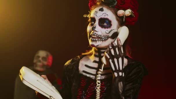 死亡女神在工作室里拿着固定电话 用绳索在办公室电话上交谈 头戴骷髅化妆 穿着圣美尔服装的女人在庆祝神圣的墨西哥仪式 手持射击 — 图库视频影像