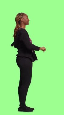 Dikey video: Afrikalı Amerikalı kız sanal görüntü ve yapay zeka kullanarak tam yeşil ekranda üç boyutlu holograma bakıyor. Tüm vücudu izole edilmiş yeşil üzerinde duran kadın modeli.