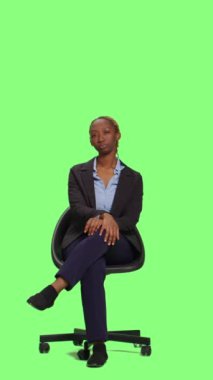 Dikey video: Şirket çalışanı sandalyede oturuyor ve stüdyoda bekliyor. Takım elbiseli genç iş kadını bekleyip hazırlanmak için yeşil perde arka planında bekliyor..