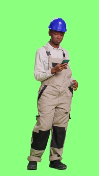 垂直视频 女建筑工人在全身绿色屏幕背景下使用智能手机应用程序 在工作室在线浏览互联网网站 用手机包工作服和硬礼帽的承包商 — 图库视频影像