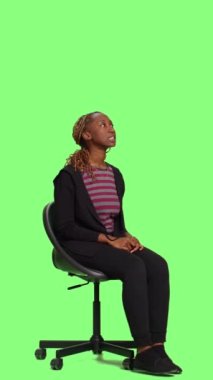 Dikey video: Meraklı mutlu kız, stüdyoda bekliyor ve hazırlanıyor, tüm vücudu yeşil ekranlı arka planda, sandalyede oturuyor. Sabırsız kadın taburede oturup bekliyor, yeşil ekran.