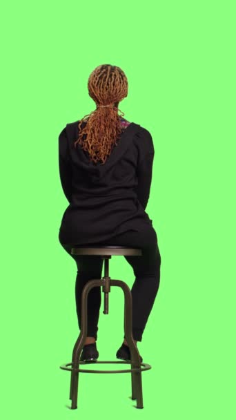 垂直ビデオ アフリカ系アメリカ人の女の子がフルボディの緑の背景の上に椅子に座って 列に並んで待って準備しています リラックスした女性は緑の画面の背景の上に準備に座っている — ストック動画