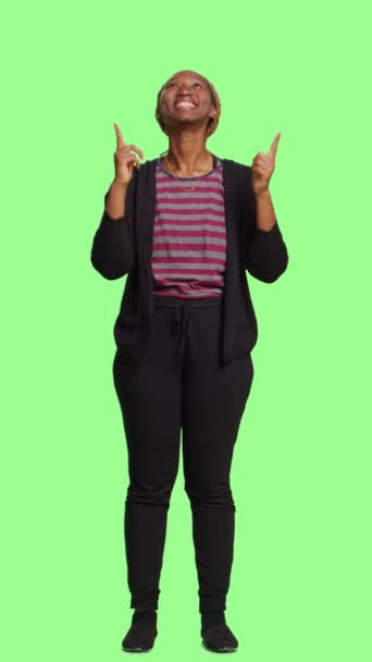 垂直视频 乐观的女性用食指指向绿色屏幕背景上的全身 指示向上的方向 年轻人推荐头上的东西 快乐的情绪 — 图库视频影像