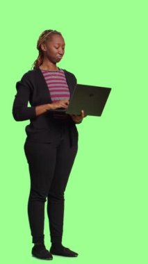 Dikey video: Bilgisayarı ve interneti kullanan Afrikalı Amerikalı kız, çevrimiçi web sitesinde gezinmek için kablosuz bilgisayar tutuyor. Gülümseyen kadın model tüm vücudun üzerinde duruyor yeşil ekran fon.