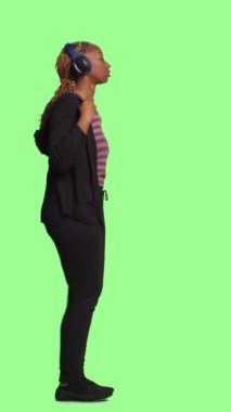 Dikey video: Genç neşeli insan kulaklıkla müzik dinliyor, tüm vücudu yeşil ekran arka planında duruyor. Kablosuz kulaklıkla mp3 şarkıları ve sesleriyle eğleniyor, neşeli davranıyor..