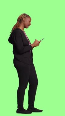 Dikey video: Cep telefonunda internette gezinen kendine güvenen güler yüzlü kız, tüm vucüt yeşil ekranında mesajlaşan kız. Çevrimiçi web sitesinde gezinmek için akıllı telefon uygulaması kullanan genç.