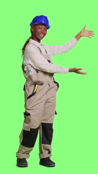 垂直ビデオ 陽気な女性コンストラクターは 広告を行うために背景の上に立って フルボディの緑の画面上に何かを広告します 背景に例を示す建設労働者は — ストック動画