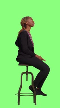 Dikey video: Genç kendine güvenen yetişkin tüm vücudu yeşil ekranda oturuyor, sırada bekliyor ve hazırlanıyor. Rahatlamış kadın yetişkin stüdyodaki yeşil ekran arka planına hazırlanıyor..