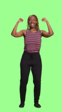 Dikey video: Afro-Amerikan modeli tüm yeşil ekran, yeşil ekran arkaplan üzerinde pozitif. İyimser bir kadın tezahürat yapıyor ve destekliyor. Stüdyo geçmişinde neşeli ve kendinden emin..