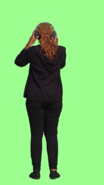 Dikey video: Neşeli modern kız kulaklıkla müzik dinliyor, arka plan tüm vücudu yeşil ekranda duruyor. Mp3 şarkıları ve kulaklıklarla eğlenmek, mutlu davranmak..