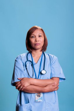 Steteskoplu yaşlı bir asistan stüdyoda kolları çaprazlanmış, mavi arka planda duruyor. Sağlıkçı üniforması giyen neşeli hemşire check-up randevusuna hazır. Tıp konsepti