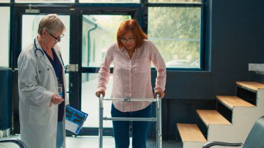 Yaşlı doktor, klinikte yürümeye çalışan, fizyoterapi randevusuna katılan yaralı hastalara destek veriyor. Bacak kırığı, ağrı ve rehabilitasyonla uğraşıyoruz..
