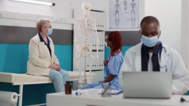 Old woman and nurse with face masks analyzing human skeleton bones at osteopathy consultation. Medical worker explaining orthopedic diagnosis on anatomy spinal cord at checkup visit.