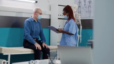 Muayenehanede eski bir hastayla görüşürken yüz maskeleri takan bir tıp çalışanı. Sağlık hizmetleri kliniğinde sağlık kontrolünde hasta bir adama danışmanlık yapan kadın hemşire..