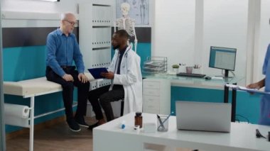 Genel pratisyen hekim, tesisteki yaşlı bir adama danışarak reçeteli ilaç ve tedavi için notlar alıyor. Sıhhiye klinikteki kıdemli hastaya muayene randevusu veriyor..