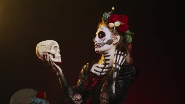 スタジオで頭蓋骨と話すラ カヴァレラ カトリーナは メキシコのハロウィンの伝統を祝うために怖いと恐怖を演じています サンタは ボディアートとホラー不気味な衣装を着ていました 手持ち撮影 — ストック動画