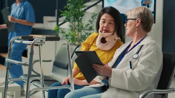 高齢の医療相談患者の首の襟のタブレットを使用してノートを取るには 待合室のロビーでの検査訪問を行う アジア人女性の身体的苦痛と傷害の予約 — ストック動画